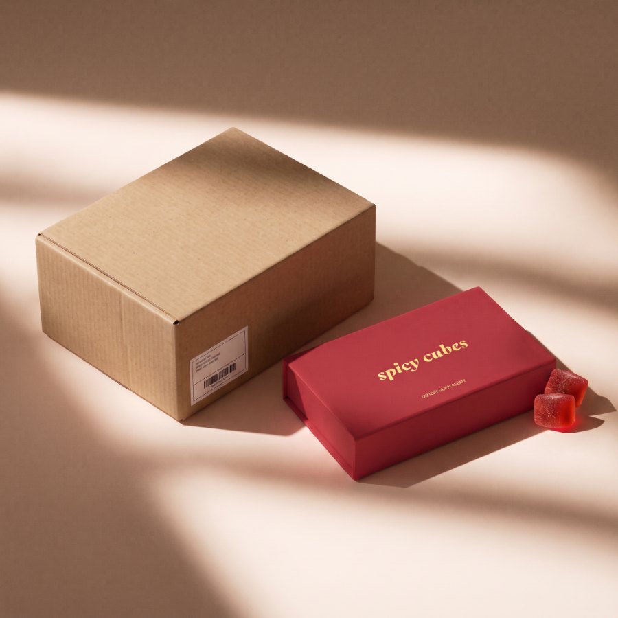 A plain unbranded kraft shipping box with only a shipping label, beside the red Spicy Cubes box with its gold foil wordmark and two cubes