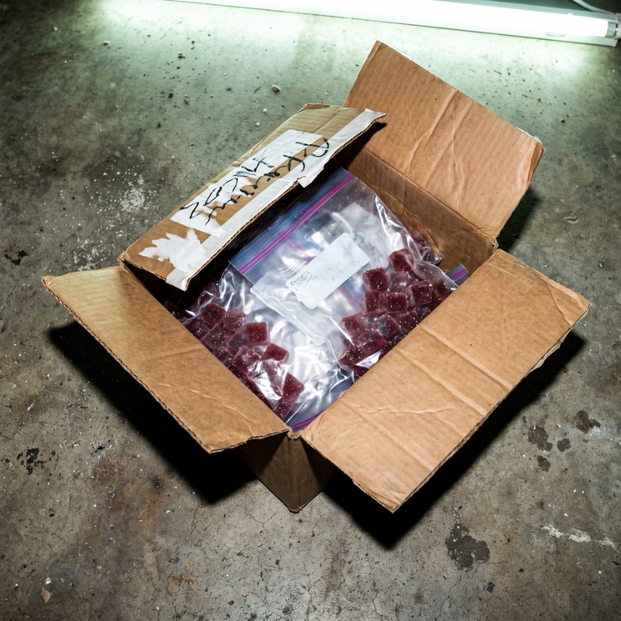 A battered cardboard box on a concrete floor holding loose gummies in an unlabelled ziplock bag